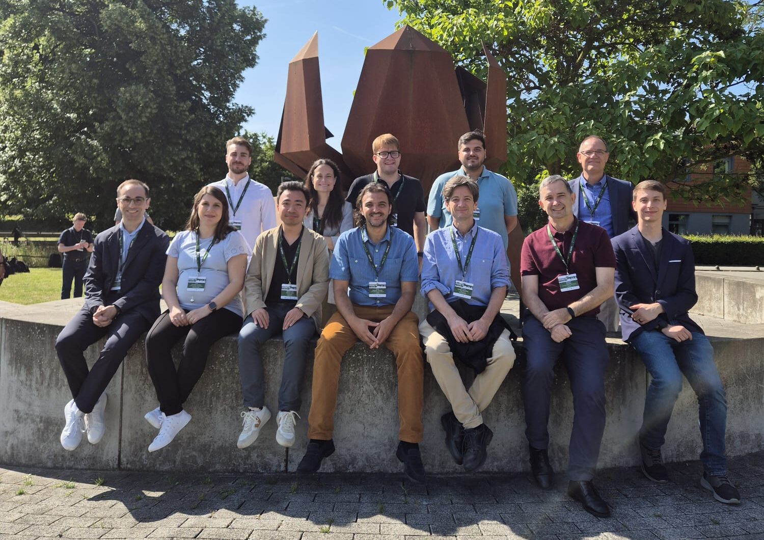 Twelve people wearing conference badges, sitting and standing in front of a rust-coloured steel sculpture